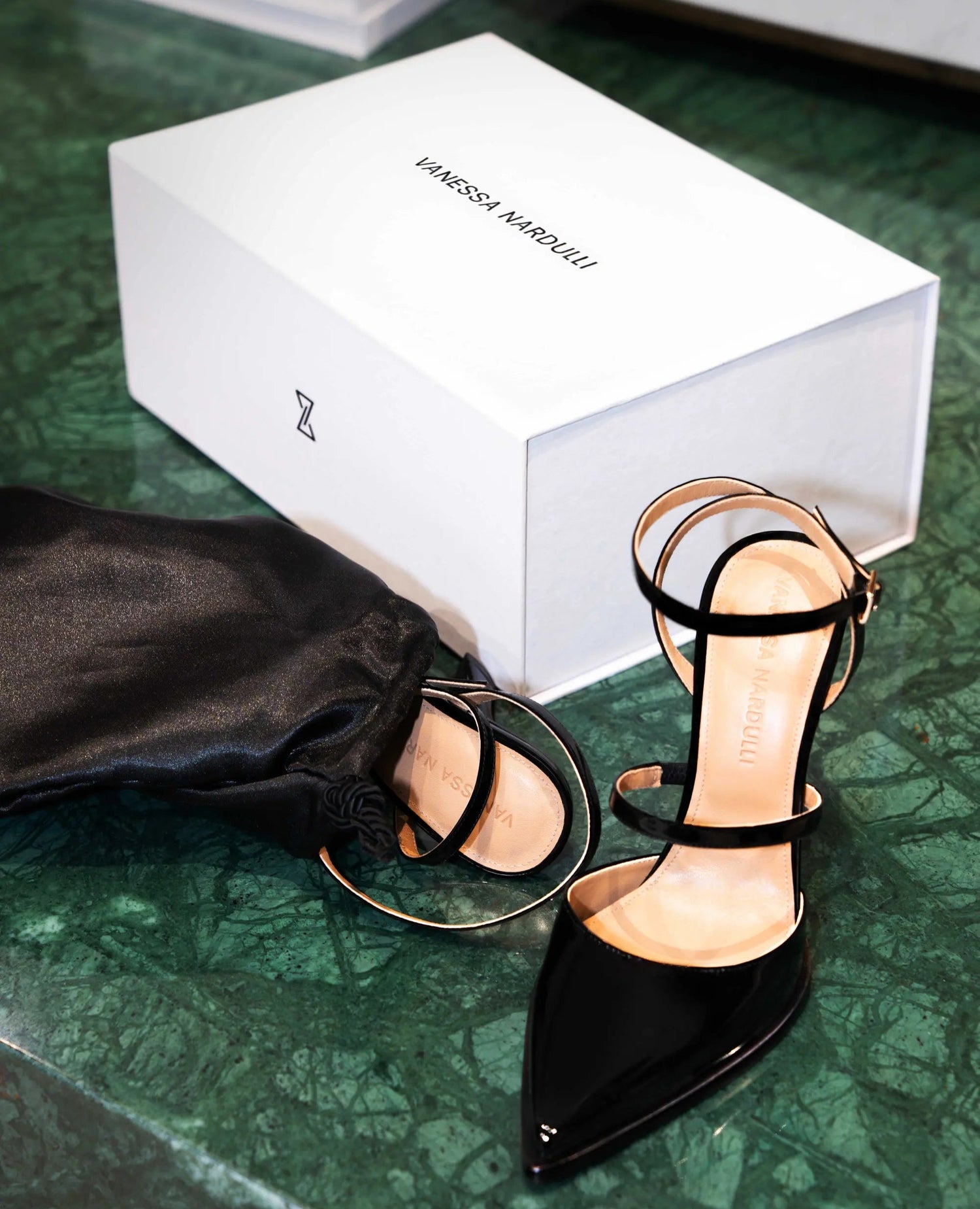 Black patent leather heels with ankle strap and geometric heel beside Vanessa Nardulli shoe box