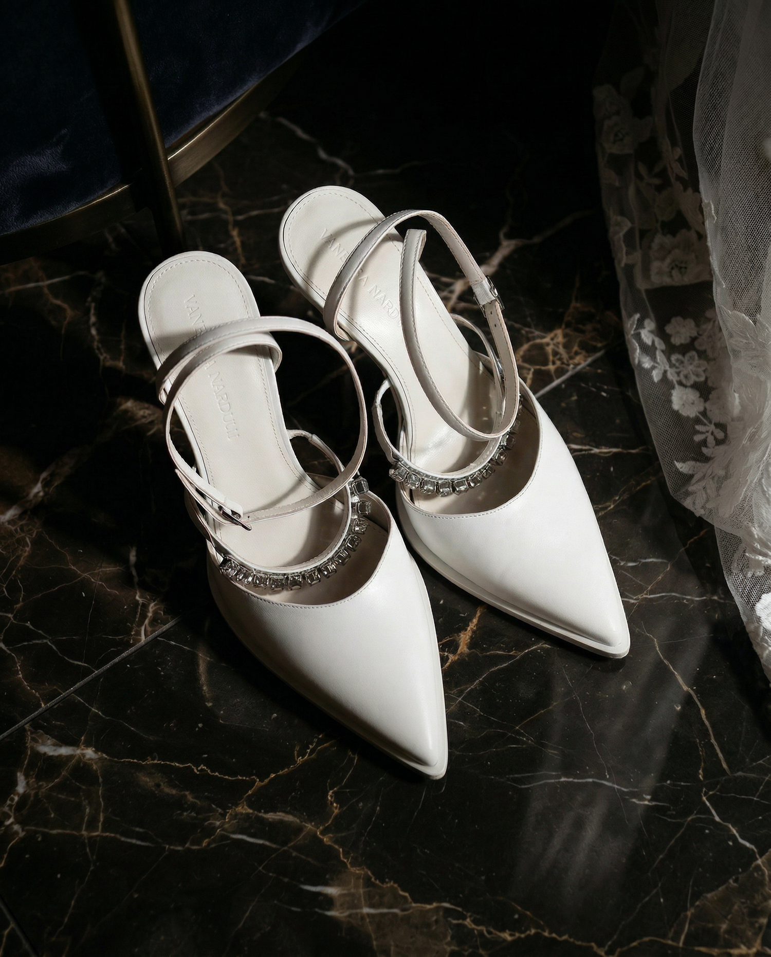White pointed-toe heels with crystal-embellished strap, displayed on marble surface